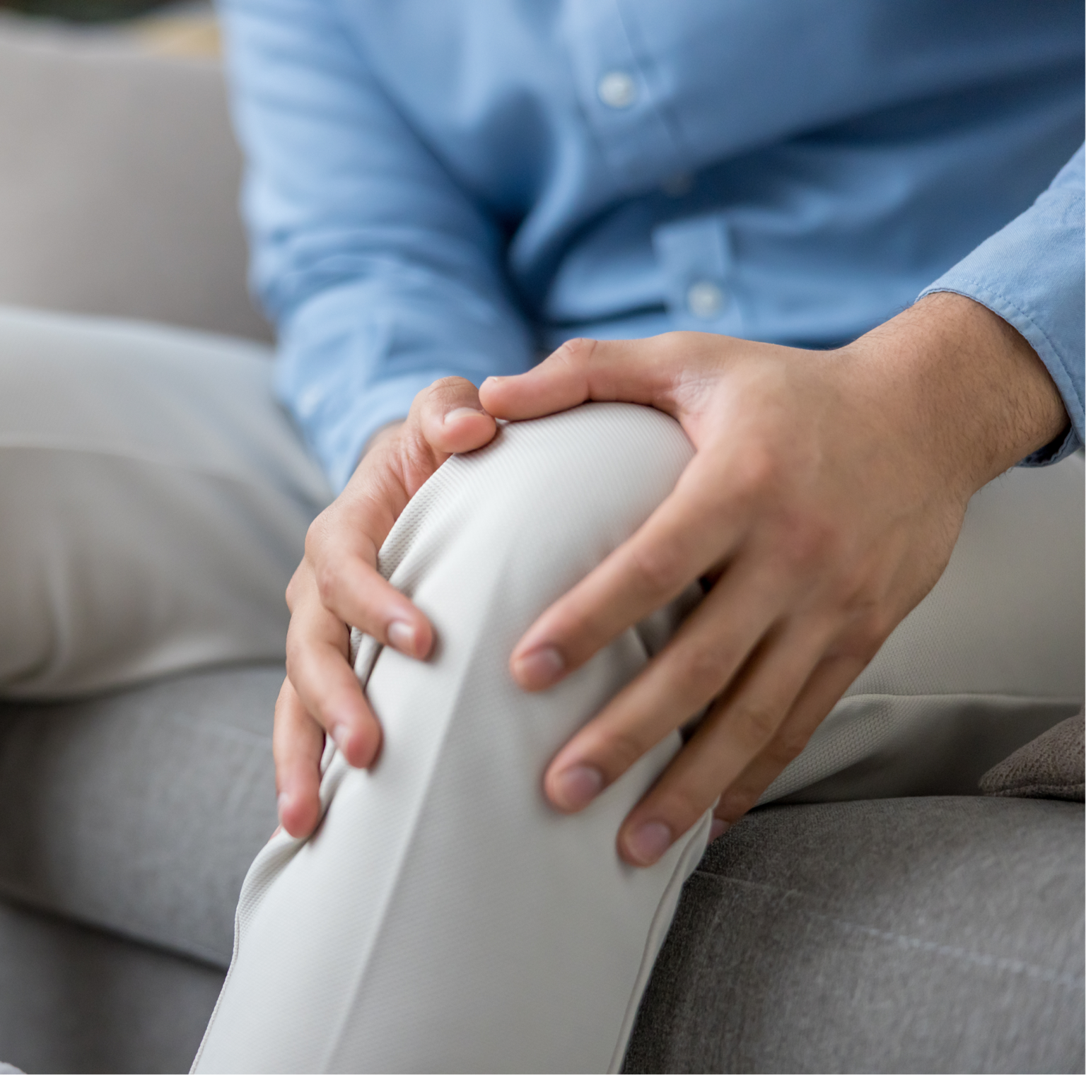 A person sitting on a couch, holding their knee in discomfort, wearing light-colored pants and a blue shirt.